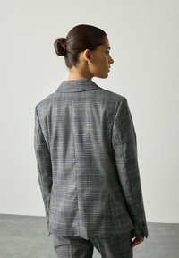 Žena s vlasy v drdolu, oblečená v šedém kostýmku s kostkovaným vzorem, stojí v minimalistické místnosti s jednoduchým světlým pozadím, otáčející se bokem.