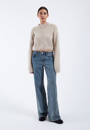 Maglione corto beige e peloso con maniche lunghe, abbinato a jeans azzurri a vita alta e scarpe nere con plateau. Sfondo semplice.