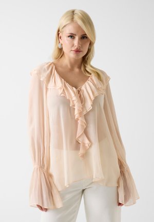 Blonde vrouw draagt een doorschijnende, licht perzikkleurige blouse met gerimpelde V-hals en uitlopende lange mouwen, gecombineerd met een witte broek tegen een effen achtergrond.