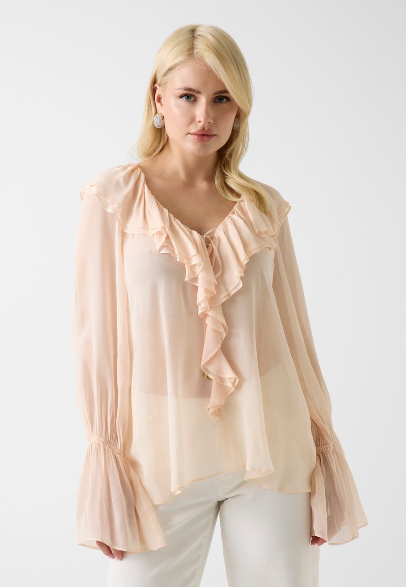 Femme blonde porte un chemisier transparent couleur pêche pâle avec col en V à volants et manches longues évasées, associé à un pantalon blanc sur un fond uni.