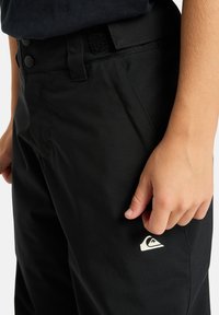 Čierne nohavice s klasickým strihom, vybavené uzatváraním na gombík a suchý zips, bočnými vreckami a bielym logom na ľavom stehne. Hladká textúra.