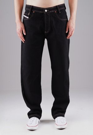 Schwarze Denim-Jeans mit lässigem Schnitt, weißen Nähakzenten, Fünf-Taschen-Design und kombiniert mit weißen Sneakers.