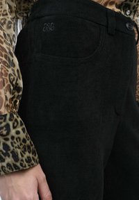 Pantalon en velours côtelé noir avec une finition texturée, doté d'une poche avant et d'une fermeture à bouton. La ceinture présente des détails de broderie discrets.
