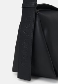 Svart läderväska med en strukturerad form, präglad logotyp på remmen och slät textur med minimalistiska designelement.