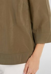 Chemise vert olive à manches trois-quarts, texture lisse, léger éclat, et ourlet incurvé, associée à un pantalon blanc.