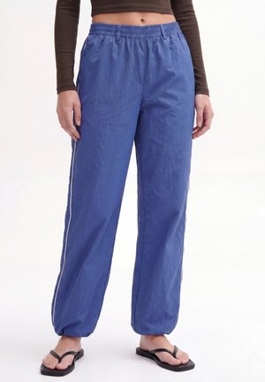 Person iført løstsiddende blå bukser med elastik i talje og ankler, sort langærmet cropped top og sorte flip-flop sandaler, stående.