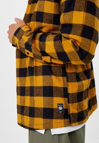 Flanelle hemd in geel en zwart ruit. Bevat knopen op de manchetten, rechte zoom en een merklabel aan de onderrand. Zachte textuur.