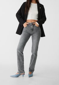 Svart kavaj över en vit ribbad crop top, kombinerad med grå raka jeans och blå spetsiga klackar, stående med händerna på höfterna.