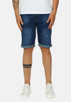 Uomo che indossa pantaloncini di denim blu scuro risvoltati, sneaker bianche e una maglietta bianca, in piedi con le braccia rilassate ai lati.