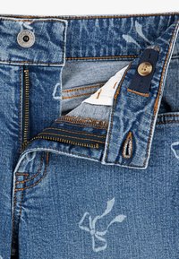 Spijkerbroek met een vervaagde blauwe kleur, gouden stiksels en een interne rits. Heeft een knoopsluiting en bloemmotieven op de stof.