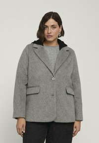 Donna con capelli corti scuri che indossa un cappotto grigio strutturato sopra un maglione grigio e pantaloni neri, su uno sfondo chiaro uniforme.