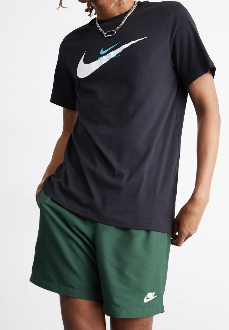Svart bomullst-shirt med en vit och teal Nike-logotyp, i kombination med gröna sportshorts med en liten vit Nike-logotyp.