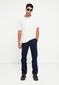 Hvid bomulds t-shirt med en blå og hvid ribbet krave, parret med mørkeblå bukser. Model iført sorte runde solbriller og brune sko.