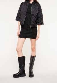 Giacca bomber nera trapuntata, vestito corto nero e stivali al ginocchio in pelle nera con finitura testurizzata e dettagli con cerniera.