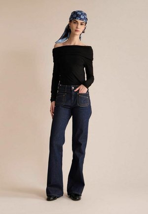 Femme portant un foulard bleu à motifs, un haut noir épaules dénudées, un jean taille haute bleu foncé et des chaussures noires, debout avec une main dans la poche.