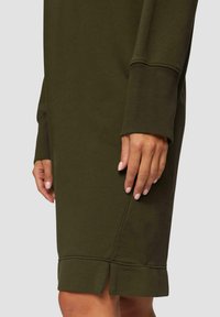 Olijfgroene sweatdress met een geribbelde manchette, een zijsplit aan de zoom en een gladde textuur. Hand rustend op de stof.