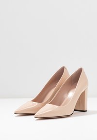 Bottines à talons hauts en cuir verni beige avec des pointes effilées et un talon large. Texture lisse avec un design élégant et un minimum de détails.