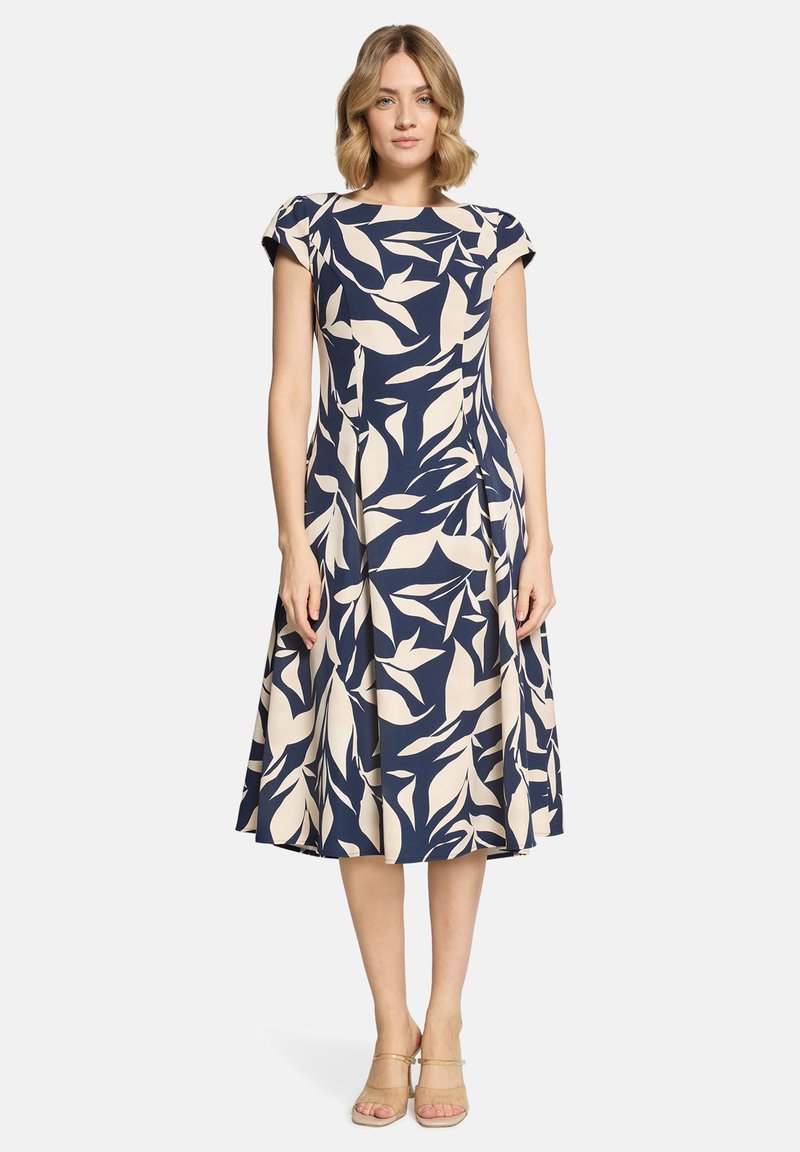 Frau trägt ein knielanges, marineblaues Kleid mit beigen, abstrakten Blattmustern und kurzen Ärmeln, steht vor einem schlichten weißen Hintergrund.