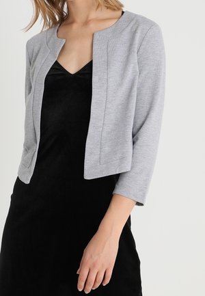 Vrouw draagt een cropped lichtgrijze blazer met open voorkant over een zwarte V-halsjurk, met één arm ontspannen langs haar zij.