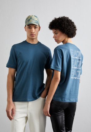 Due persone indossano t-shirt di cotone teal coordinate, con un logo sulla schiena. Una indossa pantaloni chiari; l'altra, leggings neri.