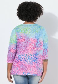 Frau mit lockigem Haar, die ein buntes, leopardgemustertes Langarmhemd und eine hellblaue Jeans trägt und vor einer weißen Wand steht.