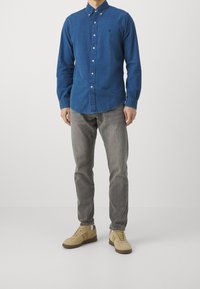 Blauwe button-up shirt met een tekstuurpatroon, gecombineerd met grijze jeans en beige sneakers. Het shirt heeft een klein logo op de borst.