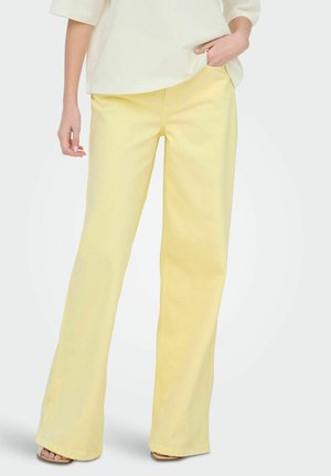 Femme portant un pantalon ample jaune clair et un chemisier crème, debout avec une main dans la poche et les pieds chaussés de sandales.