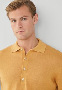 Polo shirt a maglia color senape con colletto, dotato di tre bottoni bianchi e una texture morbida.
