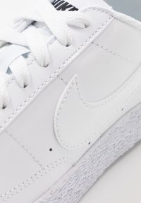 Vita lädersneaker med en texturerad sula, signaturlogotyp och platta snören. Slät yta med synliga sömmar på ovansidan.