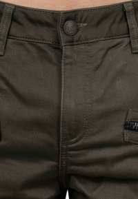 Dunkelgrüne Hose aus strukturiertem Stoff, mit einem Metallknopfverschluss und einer Seitentasche mit schwarzem Zubehör. Dezente Nähte sind sichtbar.