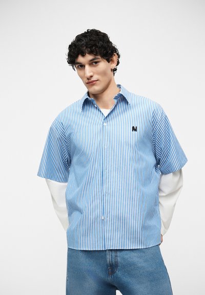 Jeune homme aux cheveux bouclés portant un tee-shirt à manches courtes rayé bleu et blanc par-dessus un tee-shirt à manches longues blanc et un jean bleu, debout.