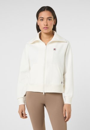 Vrouw draagt een wit jack met rits en hoge kraag met een logo op de borst, gecombineerd met lichtbruine nauwsluitende broek, staand tegen een effen achtergrond.