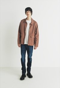 Chaqueta ligera marrón con botón en la parte delantera y dos bolsillos, llevada sobre una camisa blanca y combinada con jeans azul oscuro y botas negras.