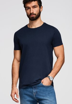 Mand med skæg iført navyblå T-shirt med korte ærmer og blå jeans, hånd i lommen, stående mod en ensfarvet lys baggrund.