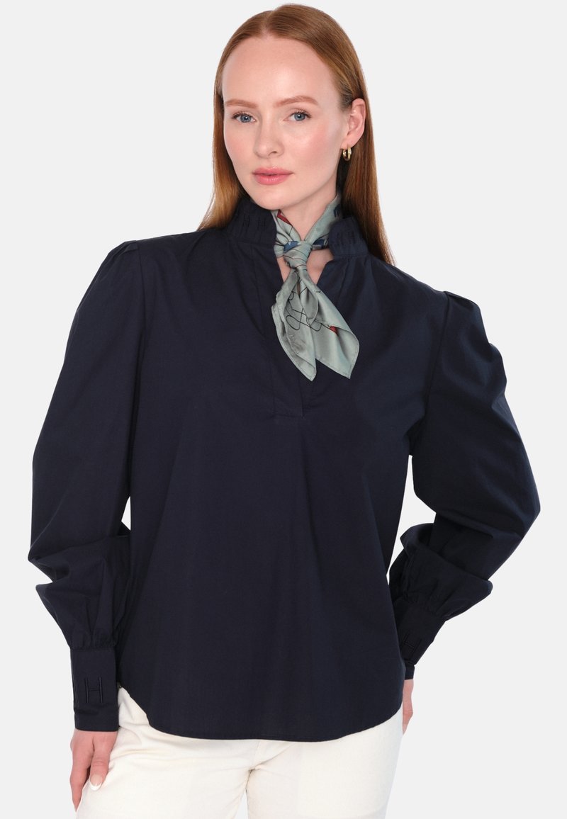 Marinebluse med høy krage, rynkedetaljer og lange ermer. Inkluderer et lysegrønt mønstrete skjerf knyttet rundt halsen.