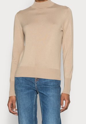 Maglione beige lavorato a maglia con collo alto, maniche lunghe e polsini e orlo ribattuti. Indossato su jeans in denim blu. Texture liscia, design aderente.