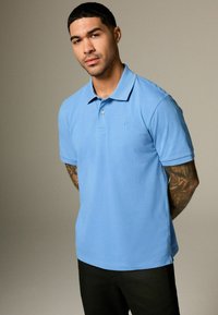 Světle modré polo triko s límečkem, krátkými rukávy a decentním logem na hrudi, spojené s tmavými kalhotami. Hladká textura.