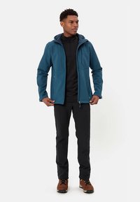 Uomo con giacca con cappuccio turchese, pantaloni neri e scarpe da trekking marroni, in piedi con le mani vicino alle tasche della giacca contro uno sfondo semplice.