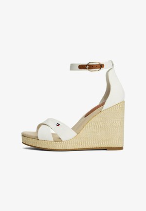 Sandales à talon compensé blanches avec une semelle jute texturée, un design à brides croisées et une bride de cheville avec une boucle en cuir marron. Petit détail de logo visible.