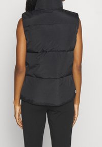 Black puffer vest with a high collar, quilted design, and a smooth texture. Features a logo detail on the lower right side.