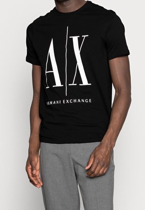 Mężczyzna noszący czarną koszulkę Armani Exchange z dużym białym logo „A|X” oraz szare spodnie, pokazany od szyi do bioder na białym tle.