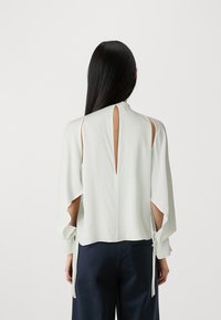 Blusa a maniche corte in tessuto verde chiaro con dettagli sulle spalle a taglio e apertura a chiave sul retro, con nastri decorativi sulle maniche.