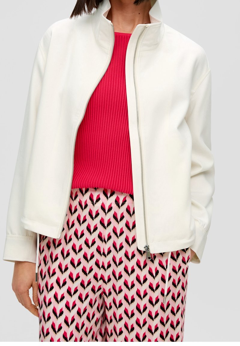Giacca bianca con zip e colletto alto, abbinata a un top rosso a costine e pantaloni rosa a fantasia. La giacca presenta una superficie liscia.
