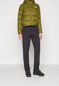 Zelená péřová bunda se zipem vpředu, kapucí a dvěma bočními kapsami, v kombinaci s černými lehkými kalhotami a černými turistickými botami.