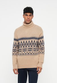 Pull en tricot beige avec un large col roulé, présentant un motif géométrique bleu foncé sur la poitrine et des manches longues avec des poignets côtelés.