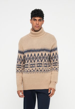 Beige gestrickter Pullover mit einem großen Rollkragen, der ein dunkelblaues geometrisches Muster auf der Brust und lange Ärmel mit gerippten Bündchen hat.