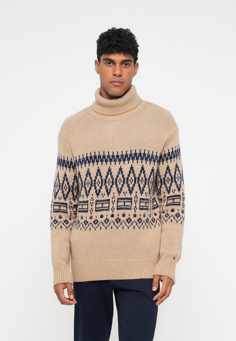 Pull en tricot beige avec un large col roulé, présentant un motif géométrique bleu foncé sur la poitrine et des manches longues avec des poignets côtelés.