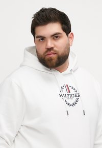 Biała bluza z kapturem z logo na przodzie ozdobionym laurami, sznurkami oraz metalowymi końcówkami. Gładka tekstura i luźny krój.