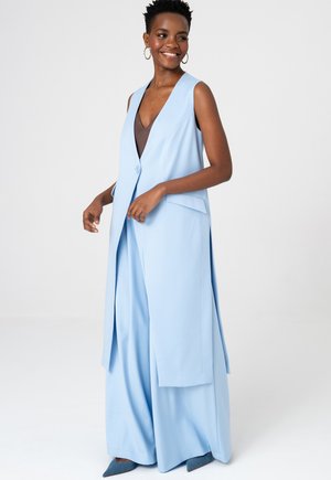 Uśmiechnięta kobieta w bezrękawnikowym, długim, jasnoniebieskim kamizelce, pasujących szerokich spodniach, brązowej bluzce i niebieskich spiczastych butach, stojąca na białym tle.