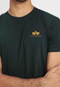 Mörkgrön bomull T-shirt med rund halsringning, med en liten orange logotyp på vänster bröst. Slät textur, korta ärmar.